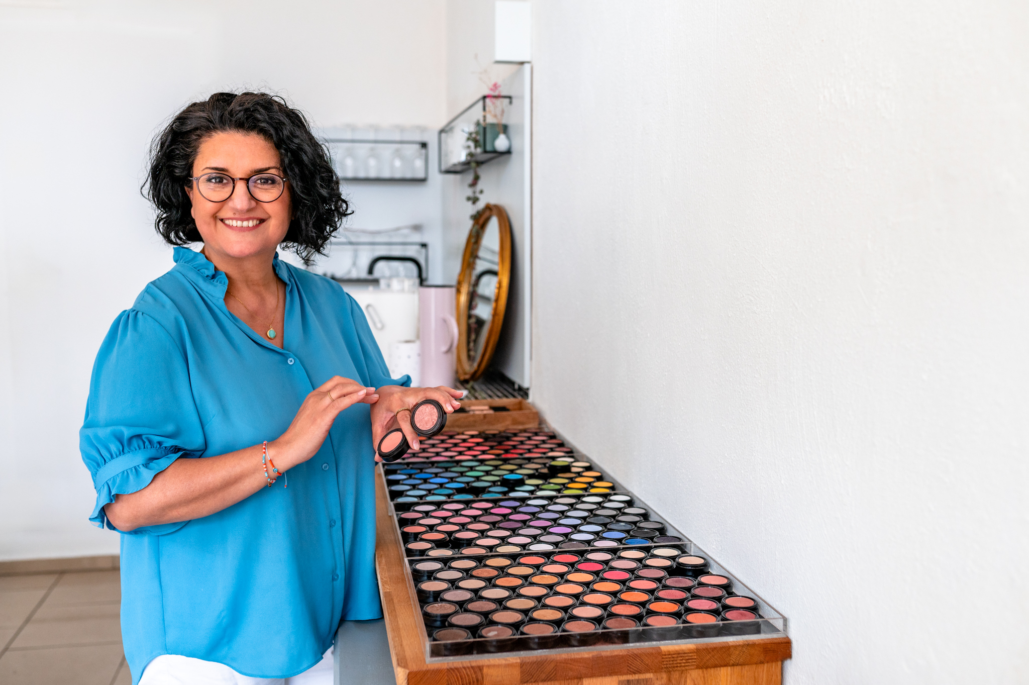Visagistin steht vor ihren Schminkutensilien und lächelt in ihrer blauen Bluse in die Kamera. Eine Expertin aus meinem Netzwerk